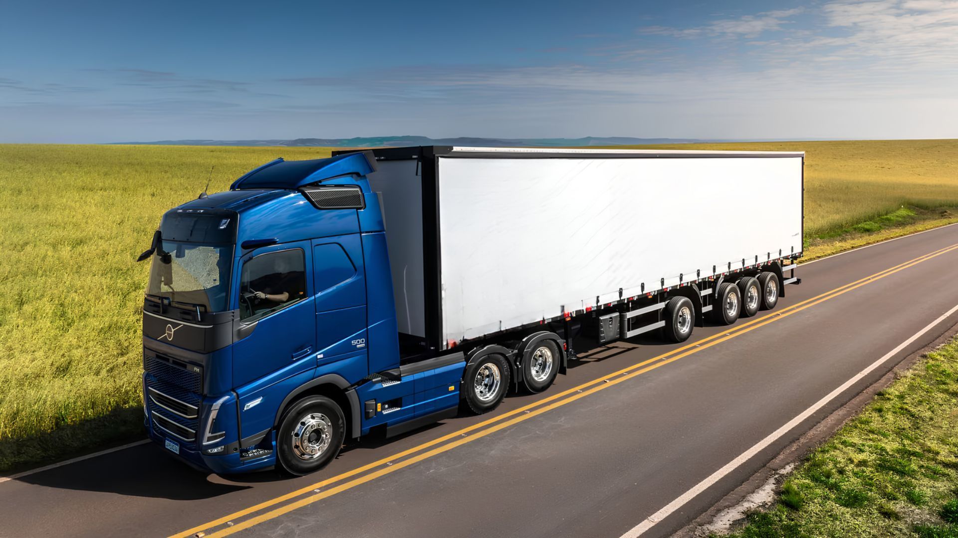 Blue semi-truck hauling a white trailer on a rural highway beside green fields
