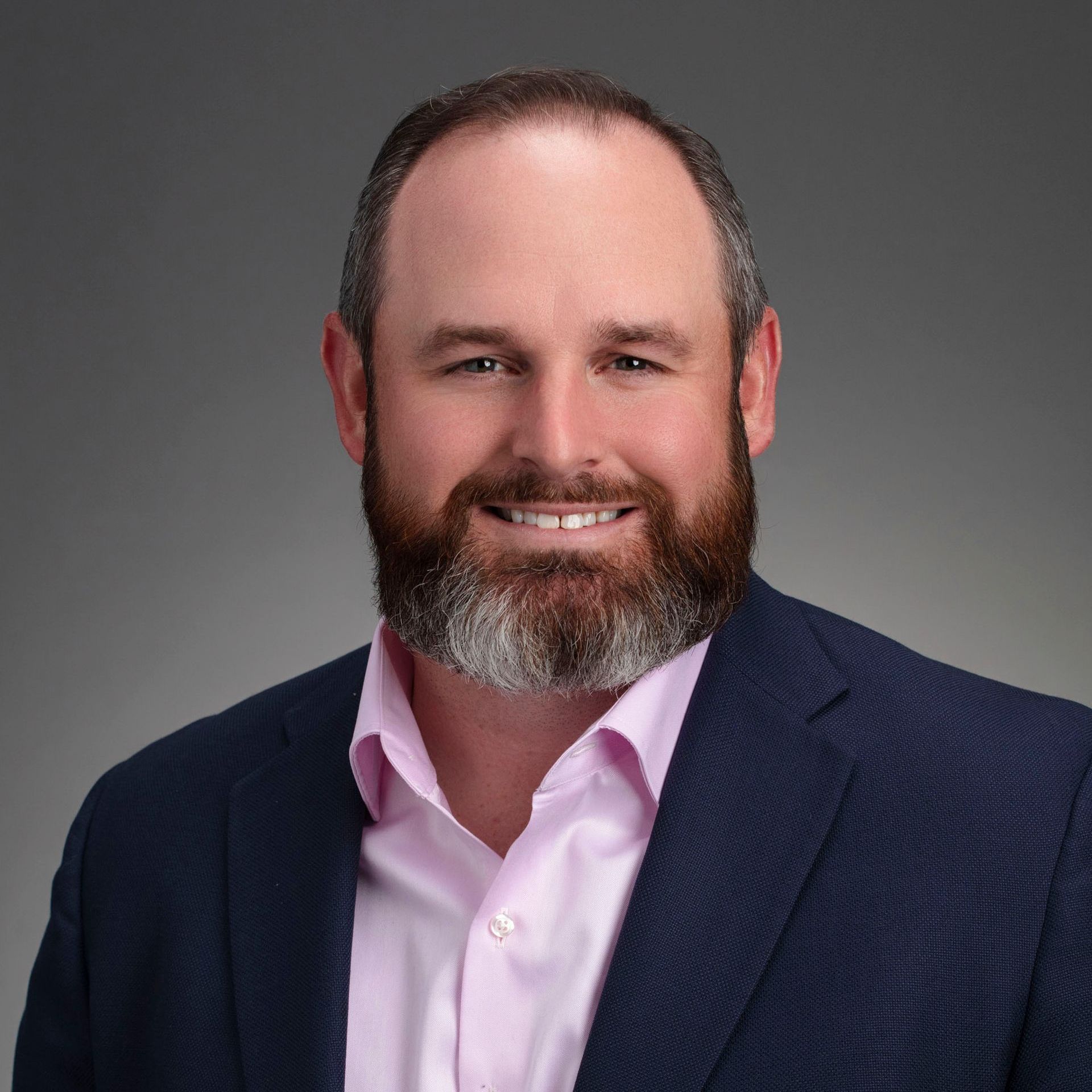 A man with a beard is wearing a blue suit and a pink shirt.