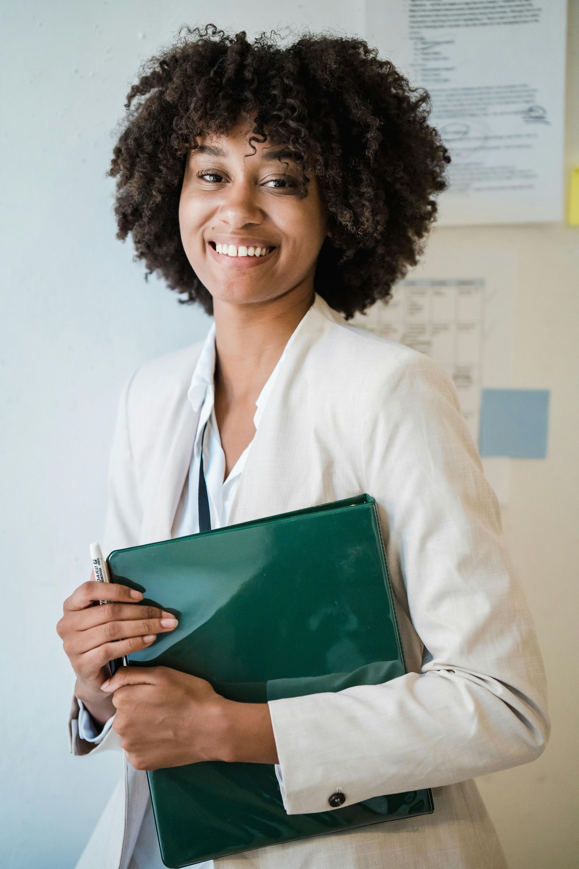 A woman in a white jacket is holding a green folder and smiling.