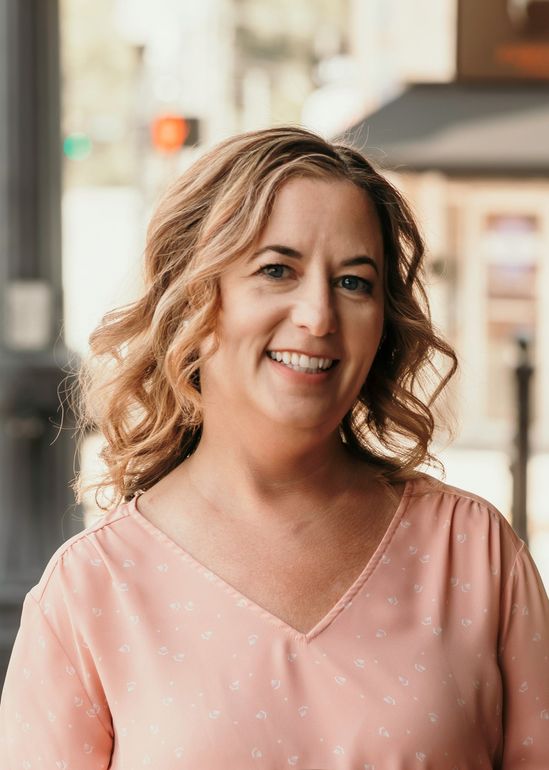 Woman with curly blonde hair smiles in a pink shirt outside a building.