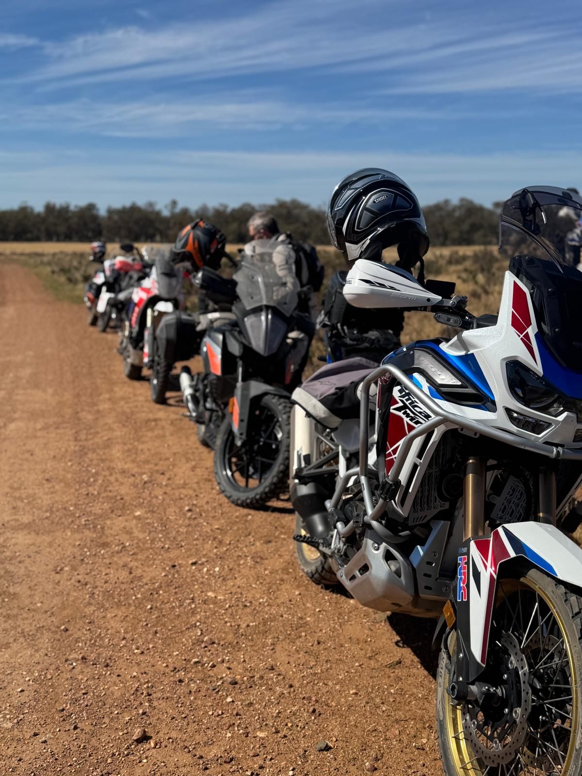 Motorcycles lined up on a dirt road under a blue sky, riders visible.