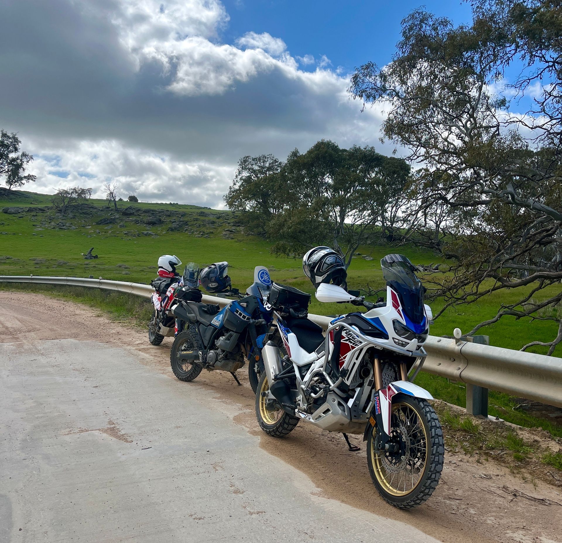 Motorcycles parked on a dirt road next to a green field under a cloudy sky.