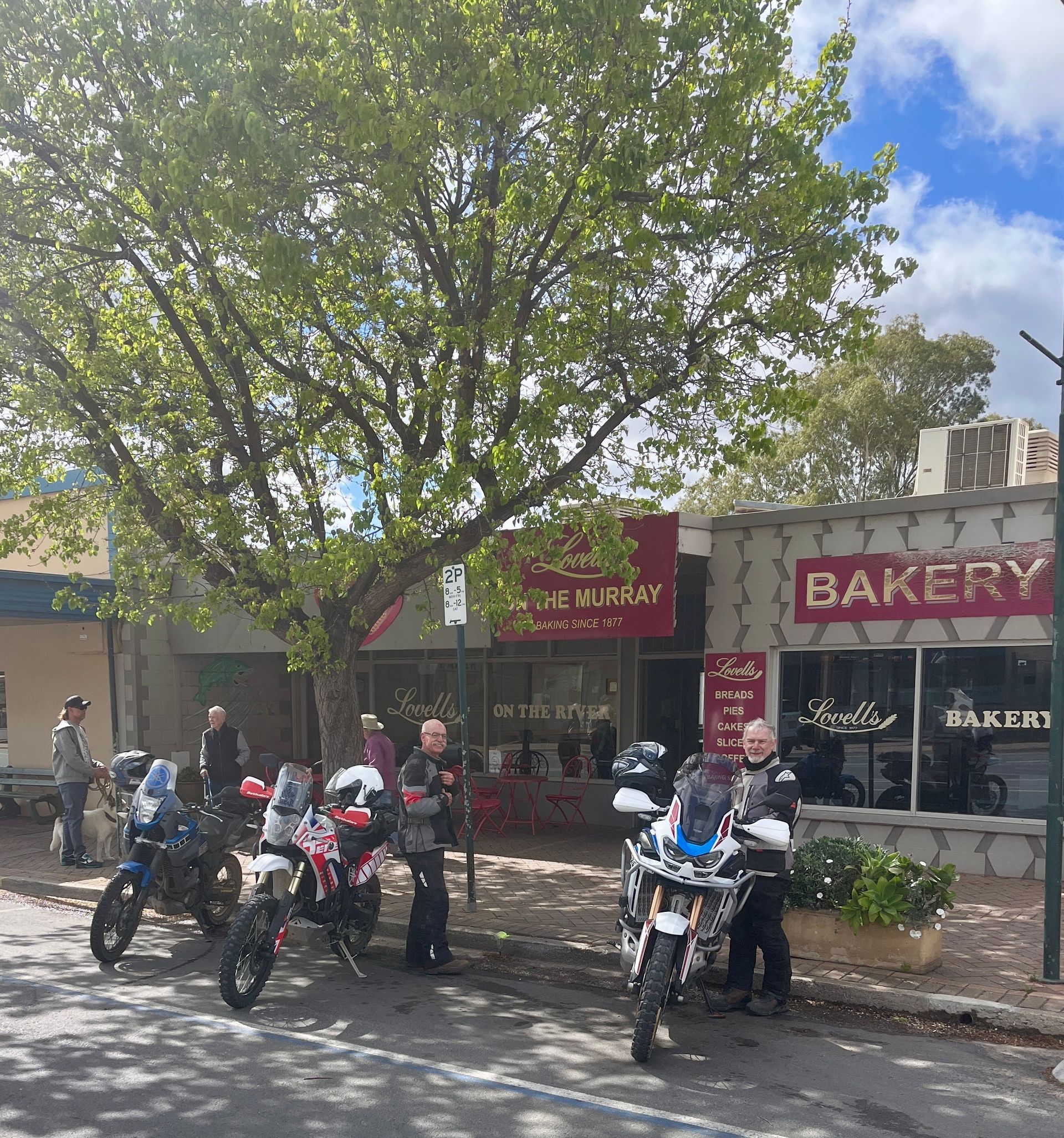 Motorcyclists outside a bakery with a red sign, under a tree, on a sunny day.