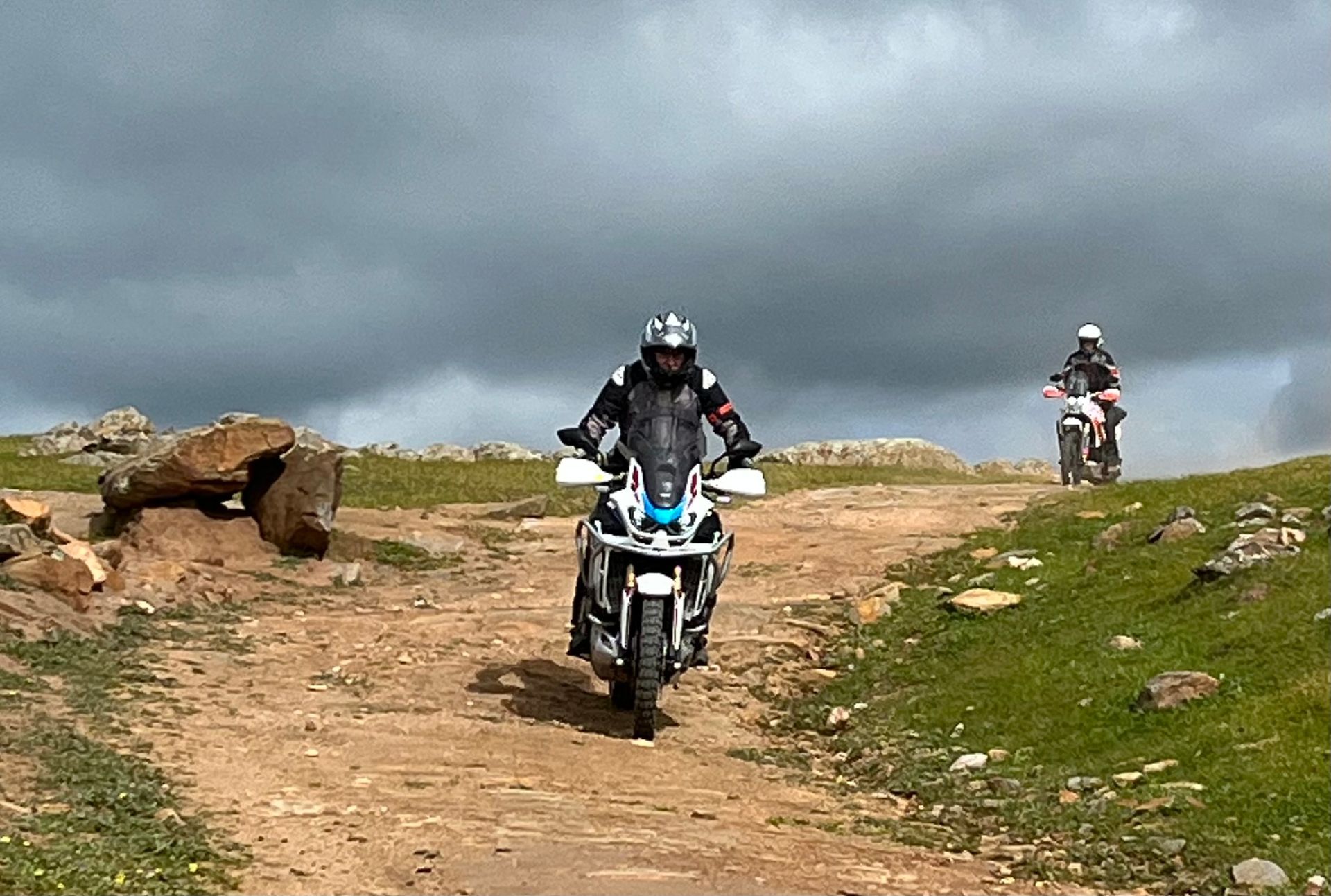 Two motorcyclists on dirt bikes riding on a rocky trail under a cloudy sky.