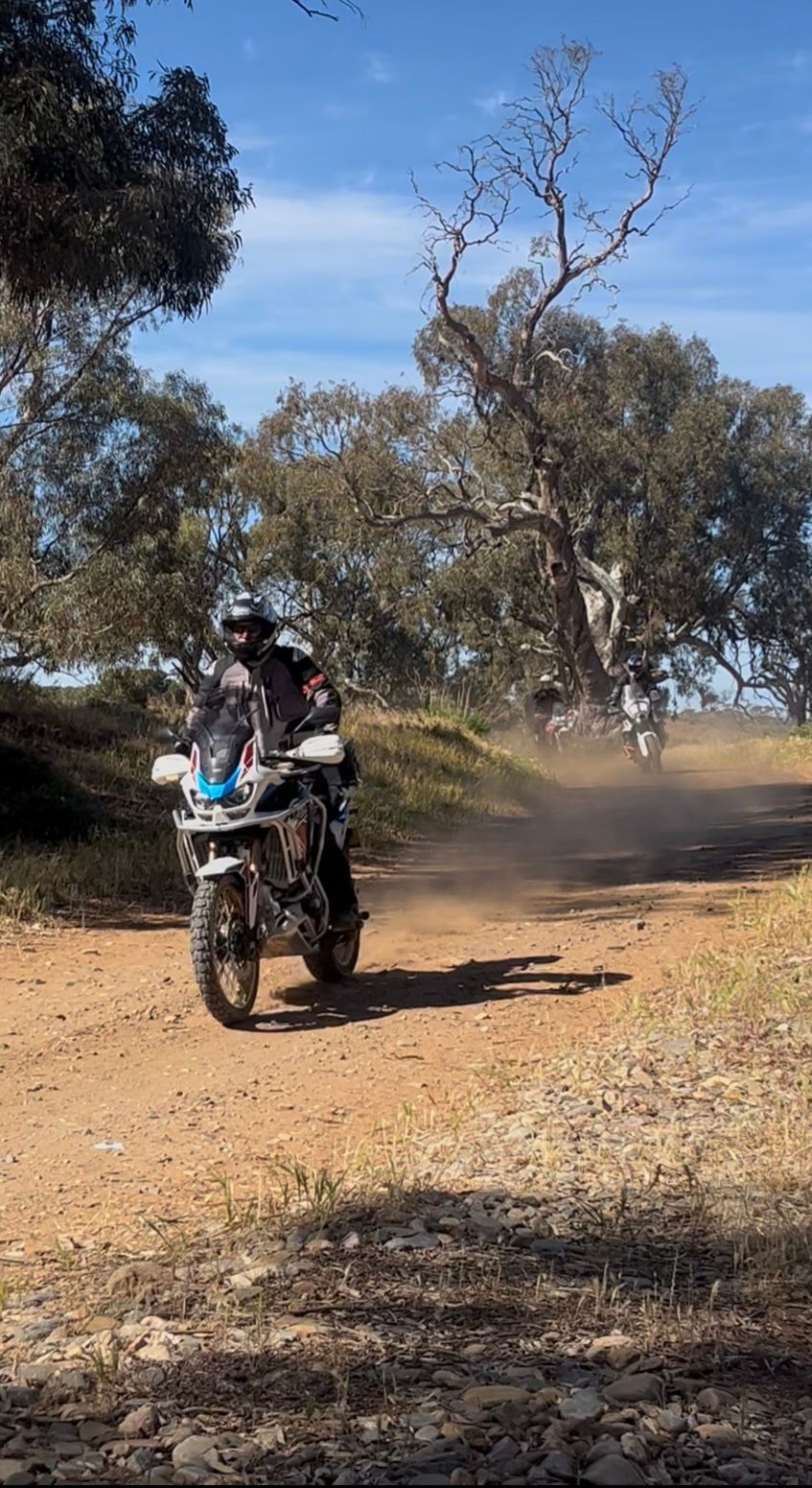 Motorcyclist riding on a dirt road, kicking up dust. A second motorcycle is visible further down the road. Sunny day.