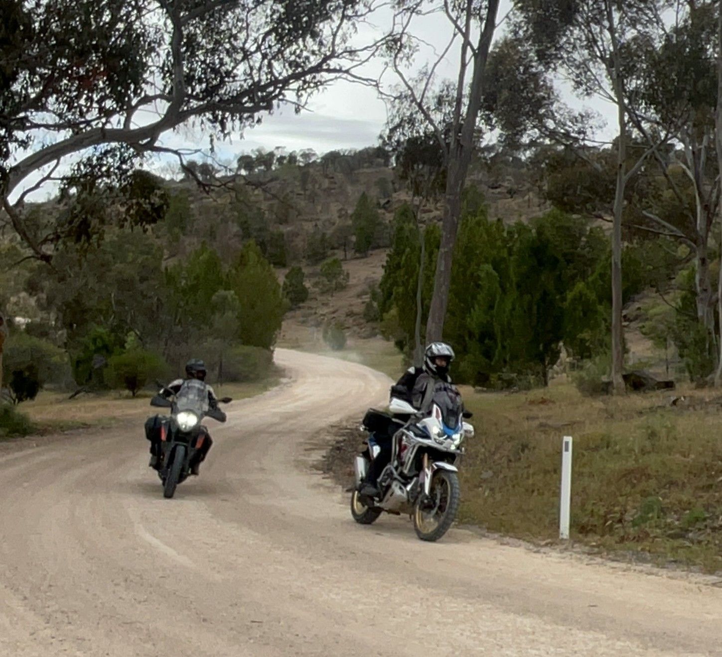 Two motorcycles riding on a dirt road through a hilly, wooded area.