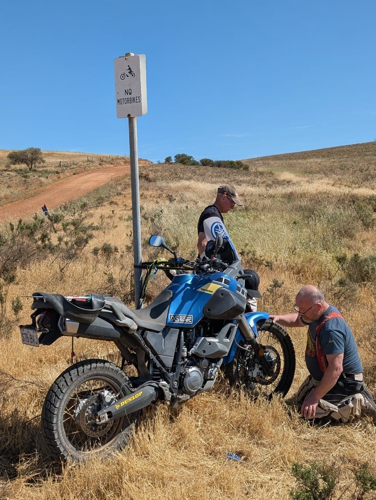 Two men near a blue motorcycle on a dirt path, one kneeling, examining the front tire. Sunny day.