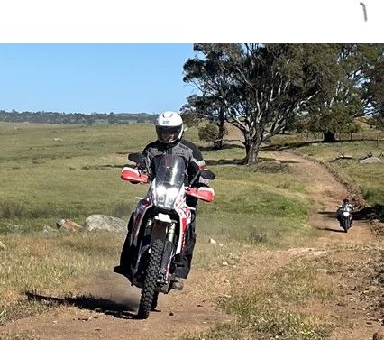 Motorcyclist riding a dirt bike on a grassy trail with another rider in the background; sunny day.