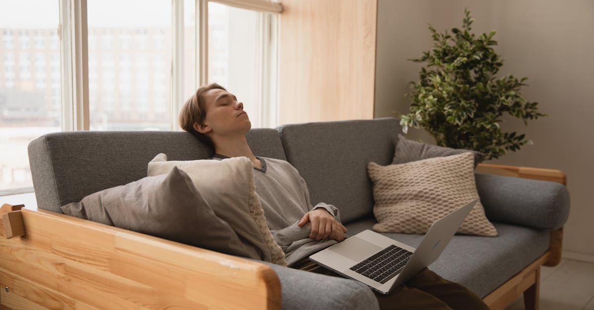 A young man sits on the couch with a laptop on his lap, exhausted with his head leaned back