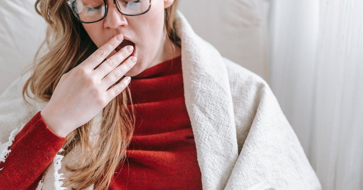 A woman in a red sweater wearing glasses is yawning, wrapped in a blanket