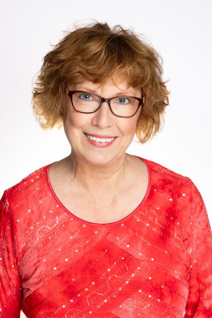 Woman with short, curly auburn hair, wearing glasses and a red patterned top, smiles at the camera.