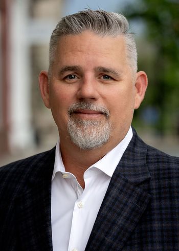 Man in blazer and open-collar shirt smiles, with salt-and-pepper hair and beard, outdoors in front of a building.