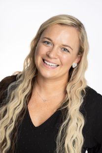 Blond woman smiling, wearing a black top and a small necklace, against a white background.