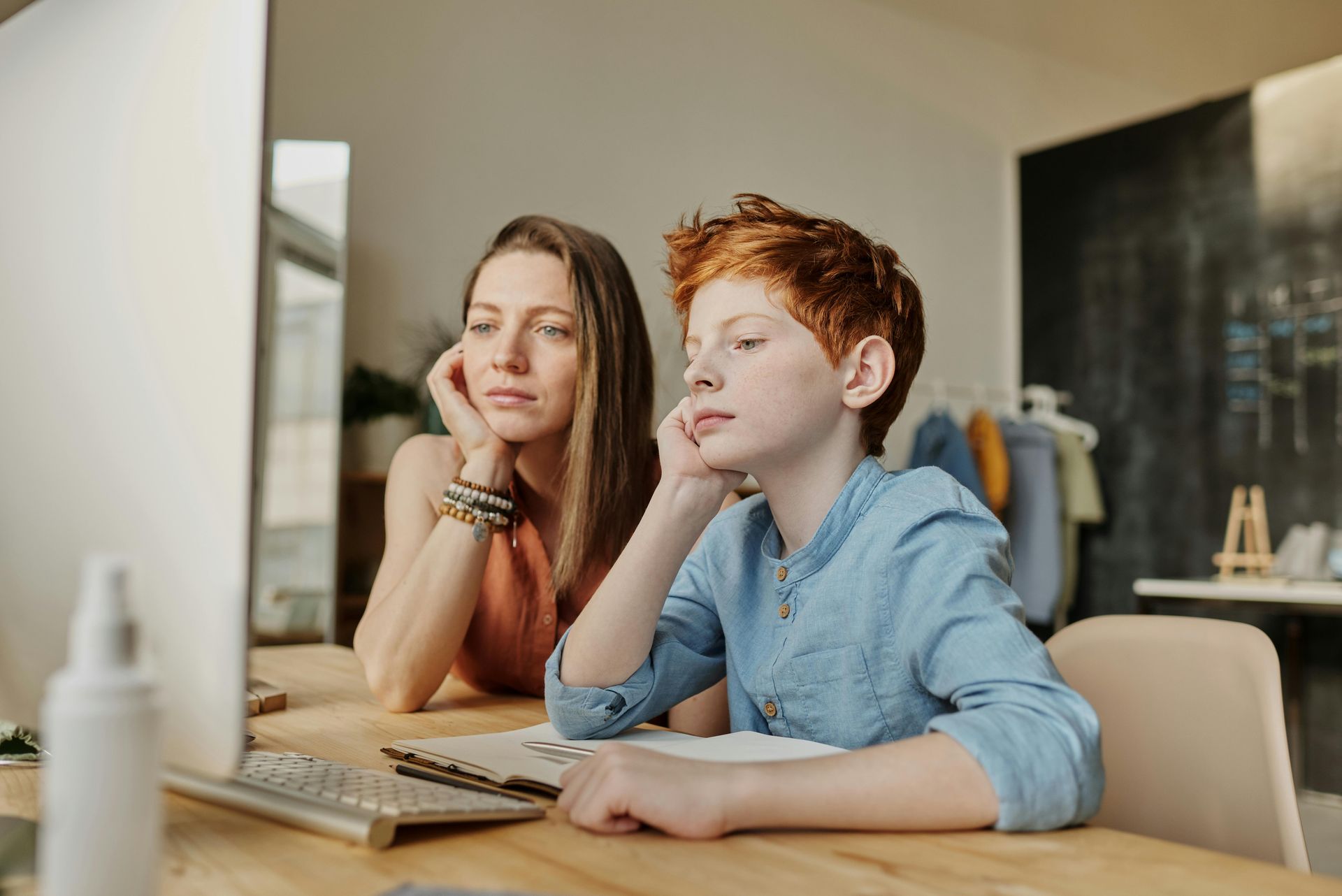 A woman and a boy are sitting at a table looking at a computer screen.