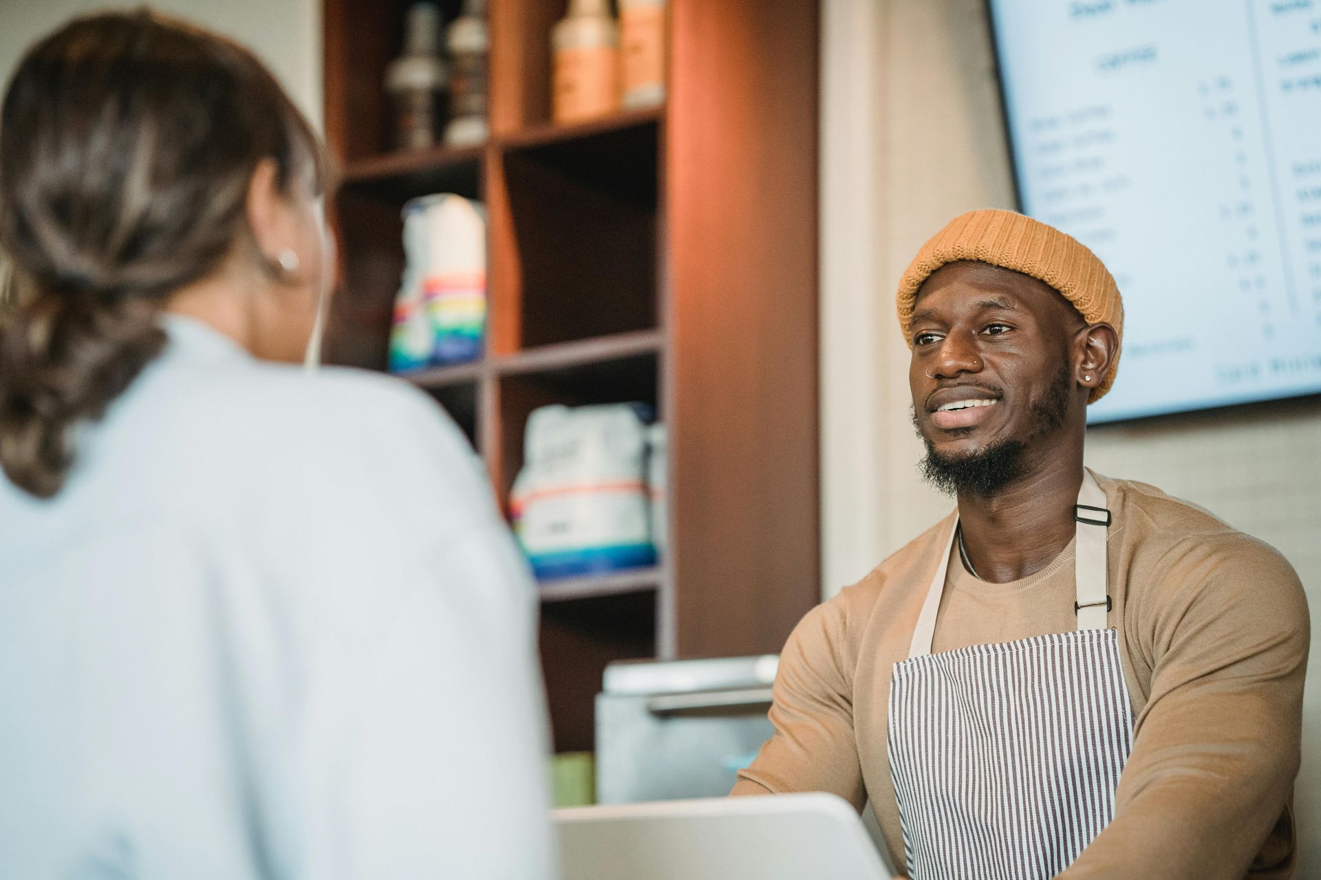 Man wearing a beanie and apron, talking to a customer behind a counter in a cafe.