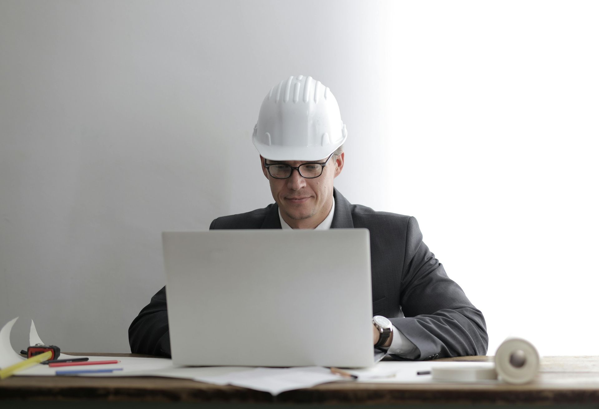 Man wearing hard hat and suit, working on a laptop at a desk with architectural plans.