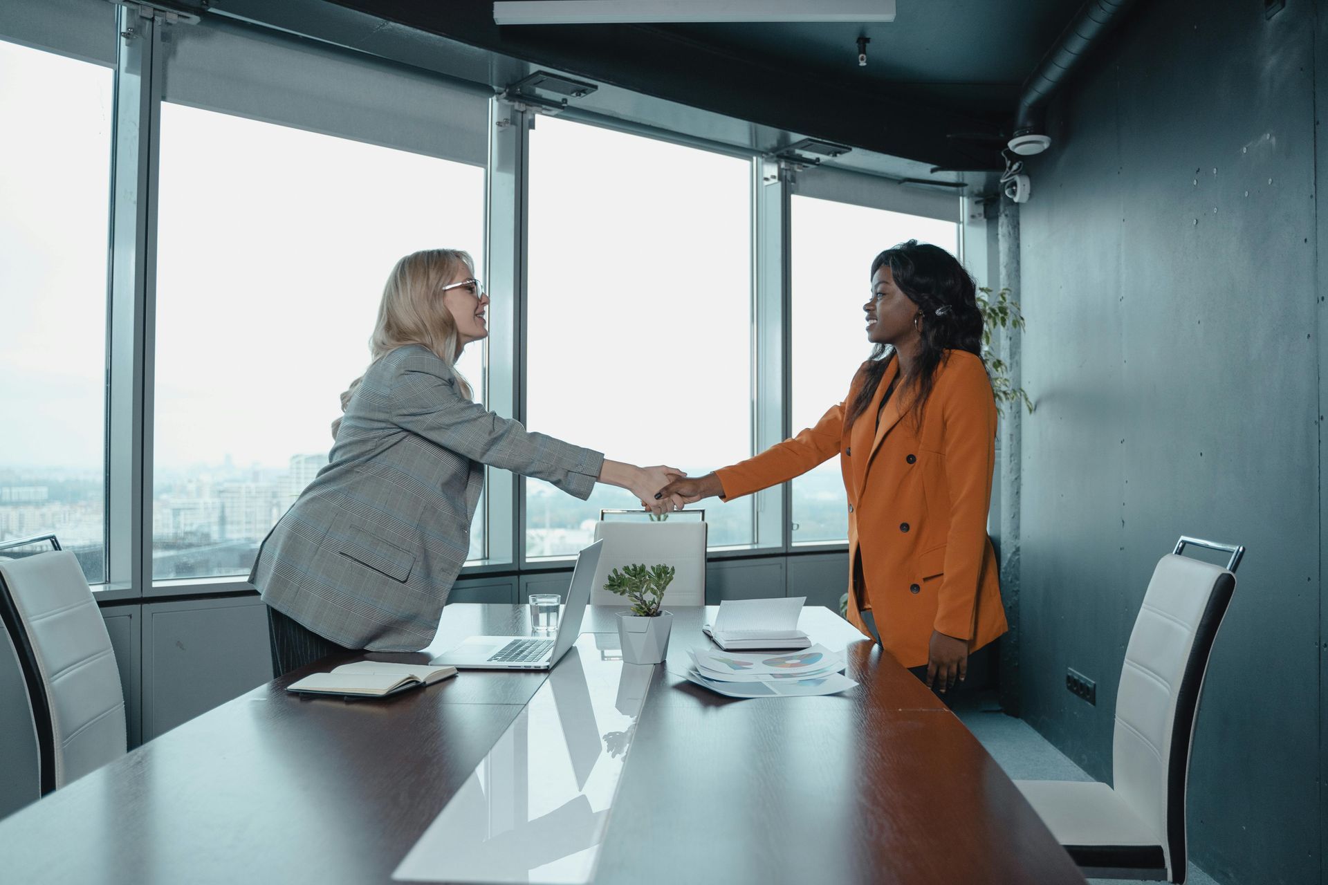 Two women shaking hands in an office, overlooking cityscape. One wears gray blazer, the other an orange blazer.