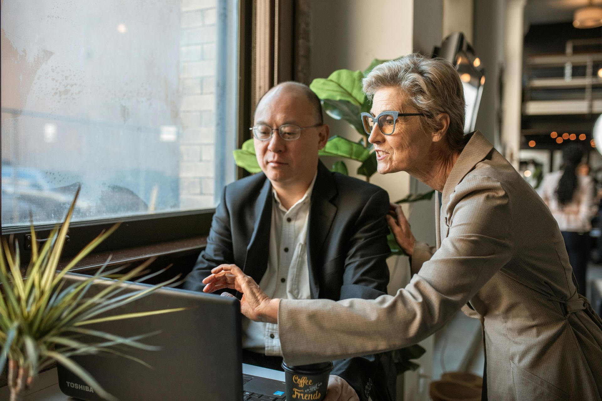 Two people looking at a laptop in a cafe setting, discussing the screen content.