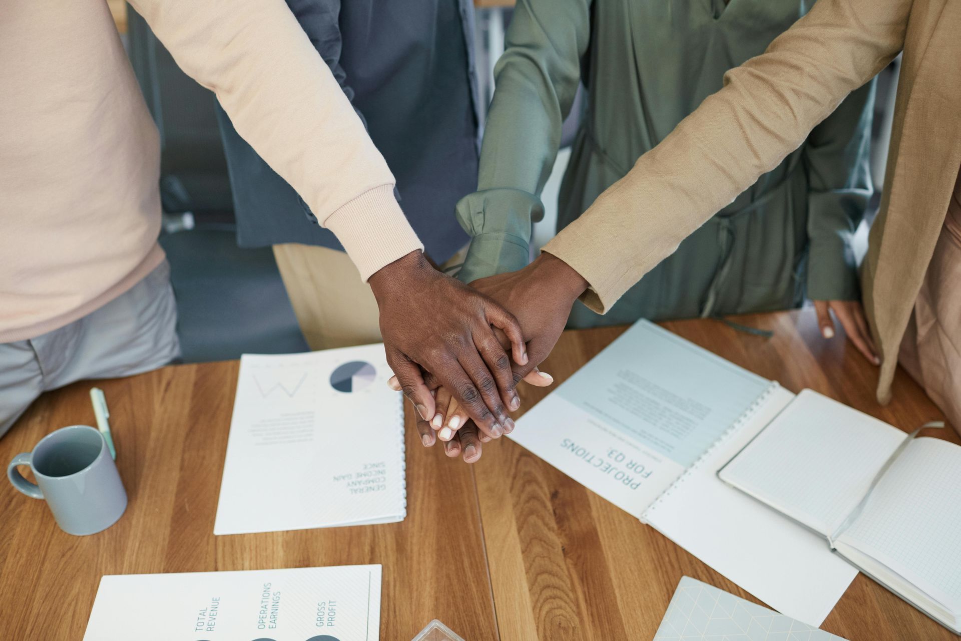 Hands of several people stacked together over documents on a wooden table.