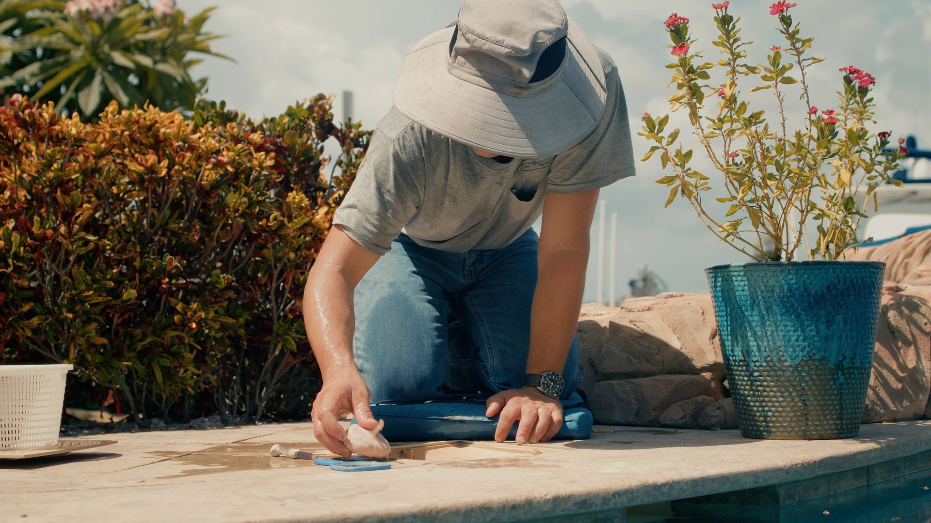 Person kneeling by pool, cleaning with water splashing. Wears hat, jeans, and a light shirt. Plant and bush in background.
