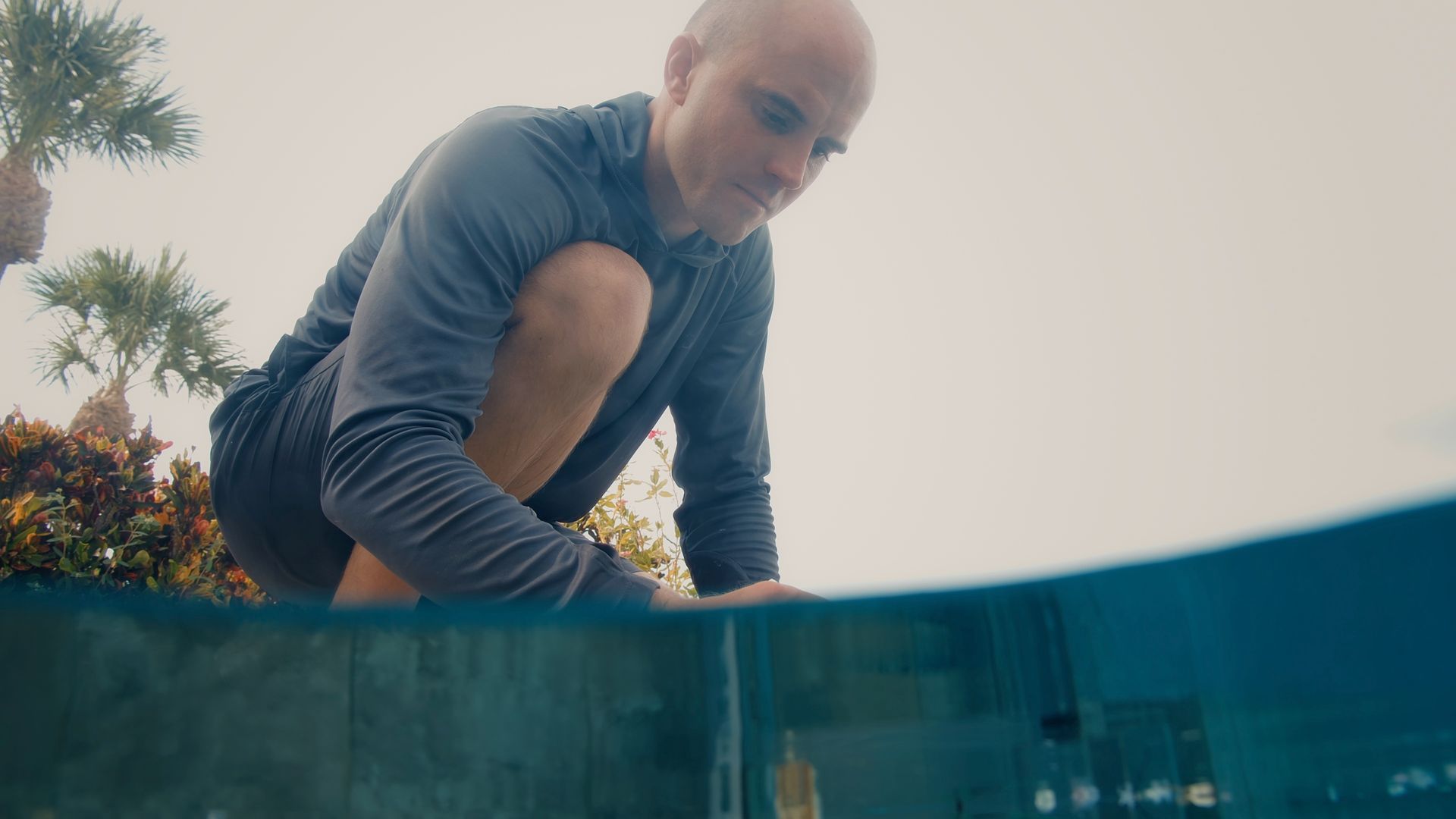 Man in athletic wear near a pool, looking down.  Half of the image is underwater.