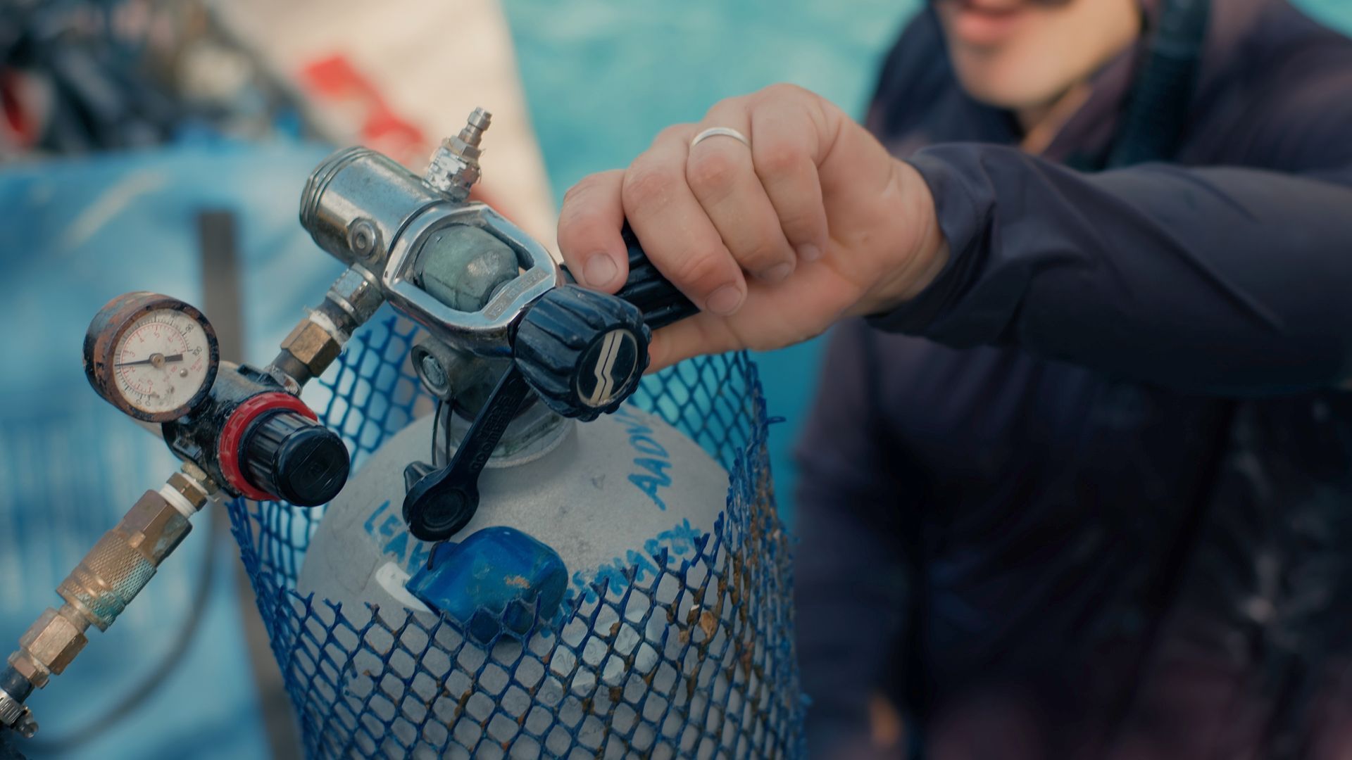 Person adjusting a scuba tank regulator with a pressure gauge. Blue tank in a net.