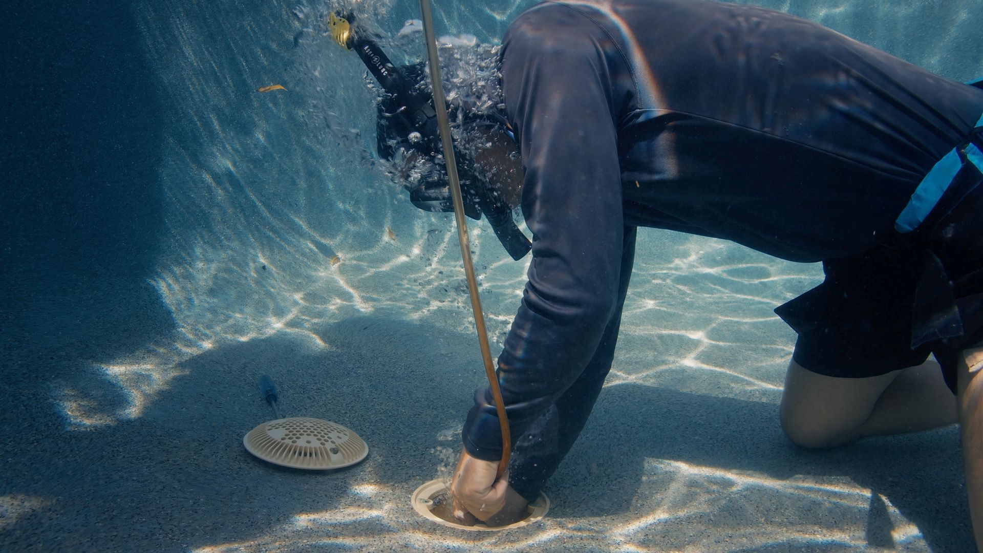 Person underwater wearing snorkel, reaching into hole in sand next to a flat disk.