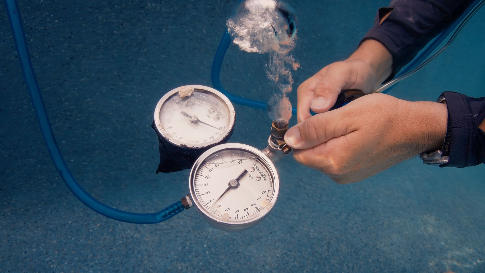 Diver checking gauges underwater; blue water, two white-faced pressure gauges, hands.