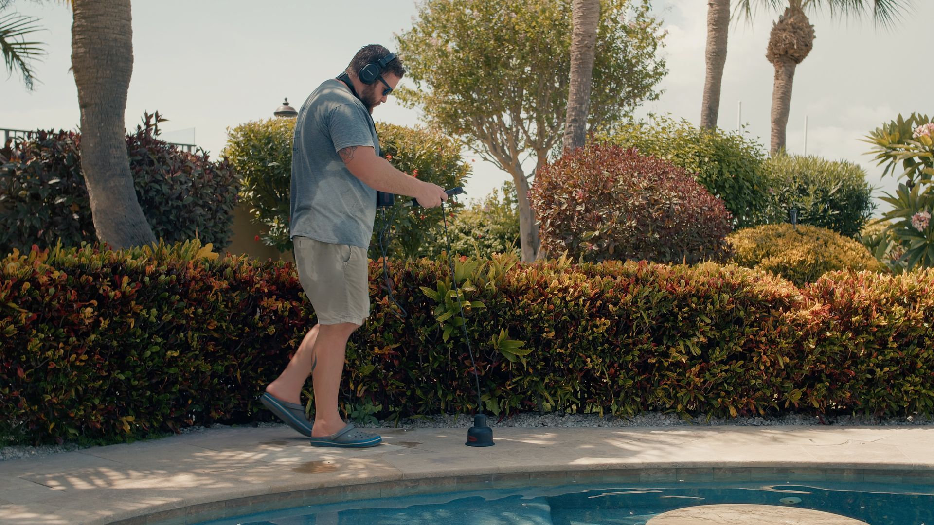 Man in shorts near pool, dropping object into water. Sunny outdoors with bushes and trees.