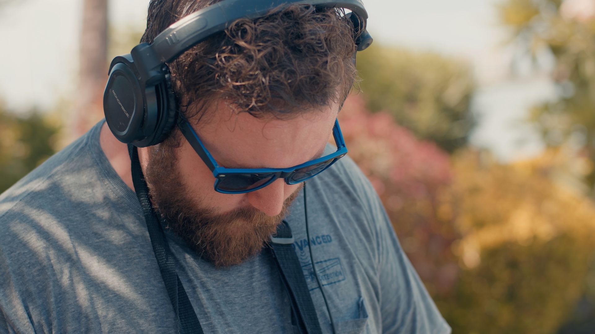 Man with beard, wearing sunglasses and headphones outdoors, looking down.