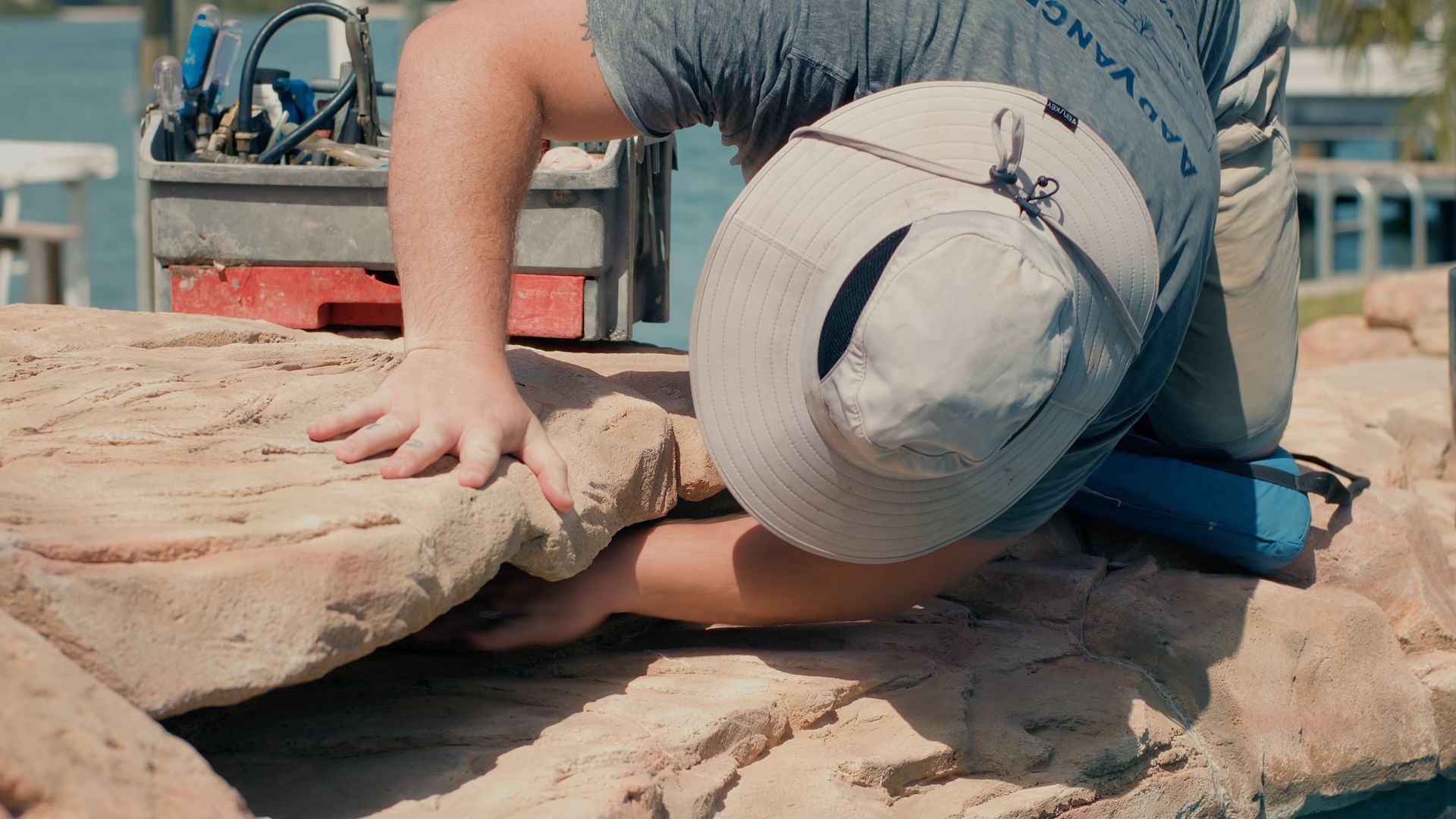 Person in gray shirt and sun hat, working on a rock structure.