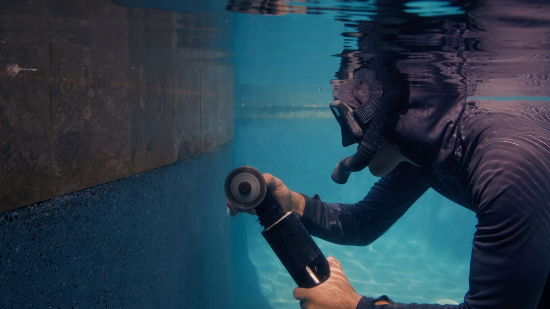 Scuba diver welding underwater on a tiled wall, wearing a black wetsuit and using an underwater welding torch.