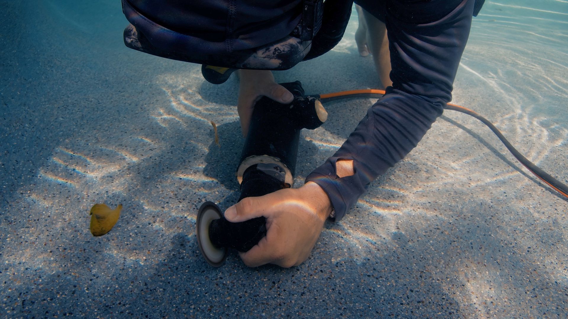 Person underwater working on black equipment in a pool.
