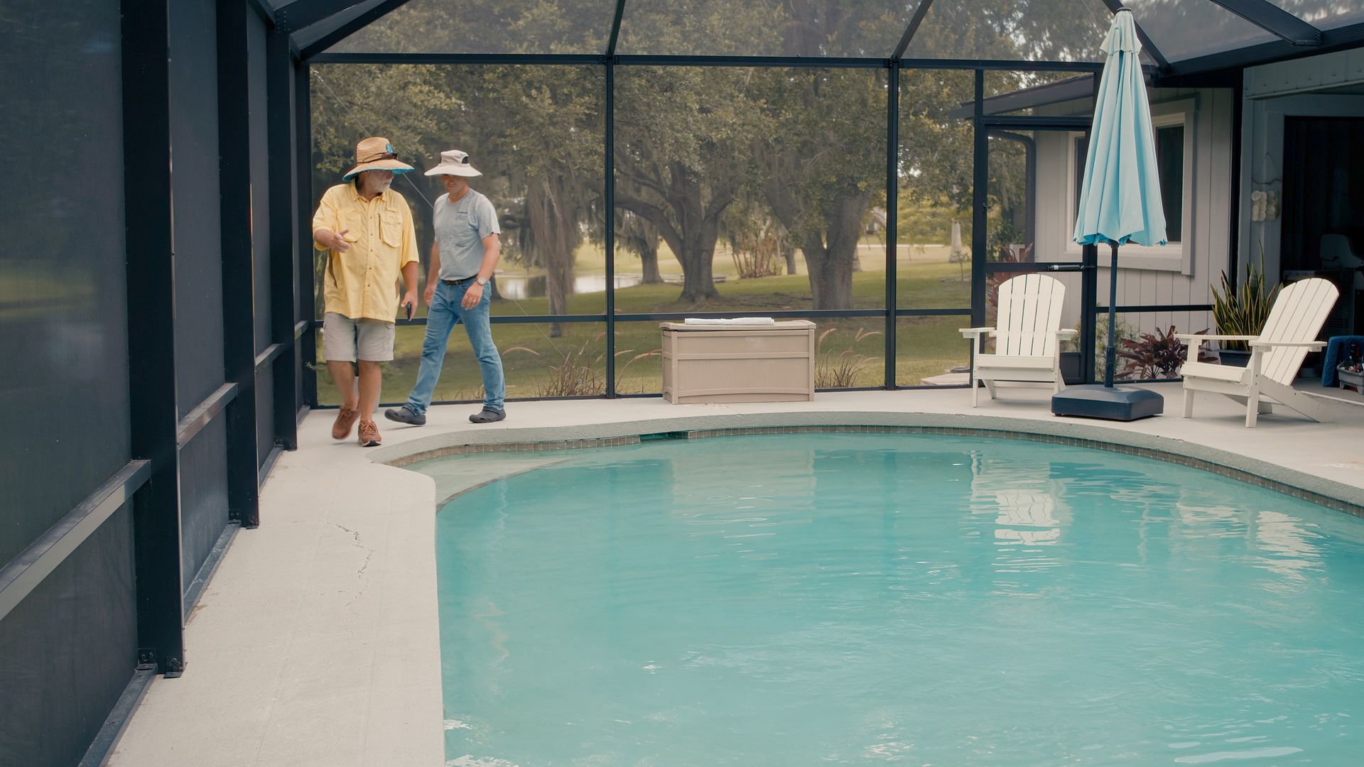 Two people wearing hats walk beside a pool enclosed by a screen, trees visible in the background.