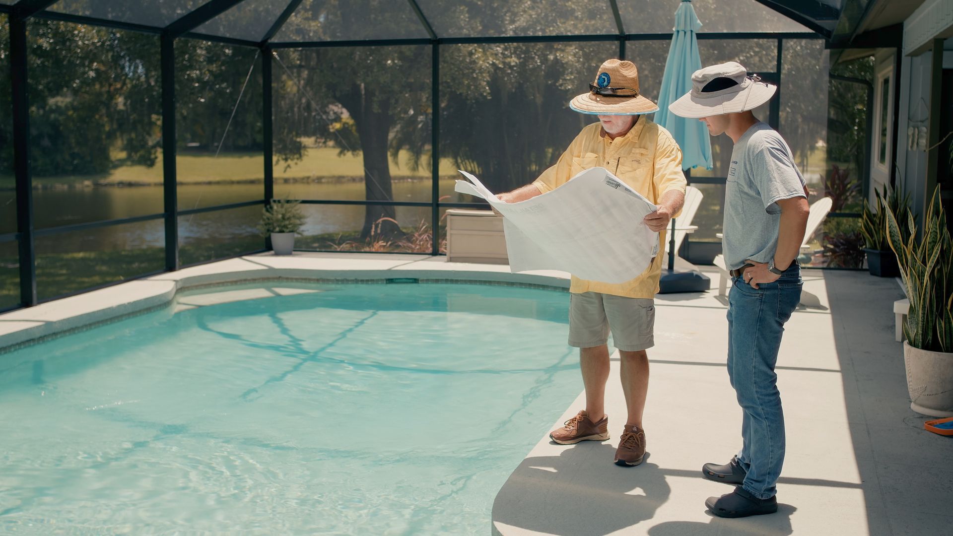 Two men by a swimming pool, examining a white pool cover. One man points to the cover.
