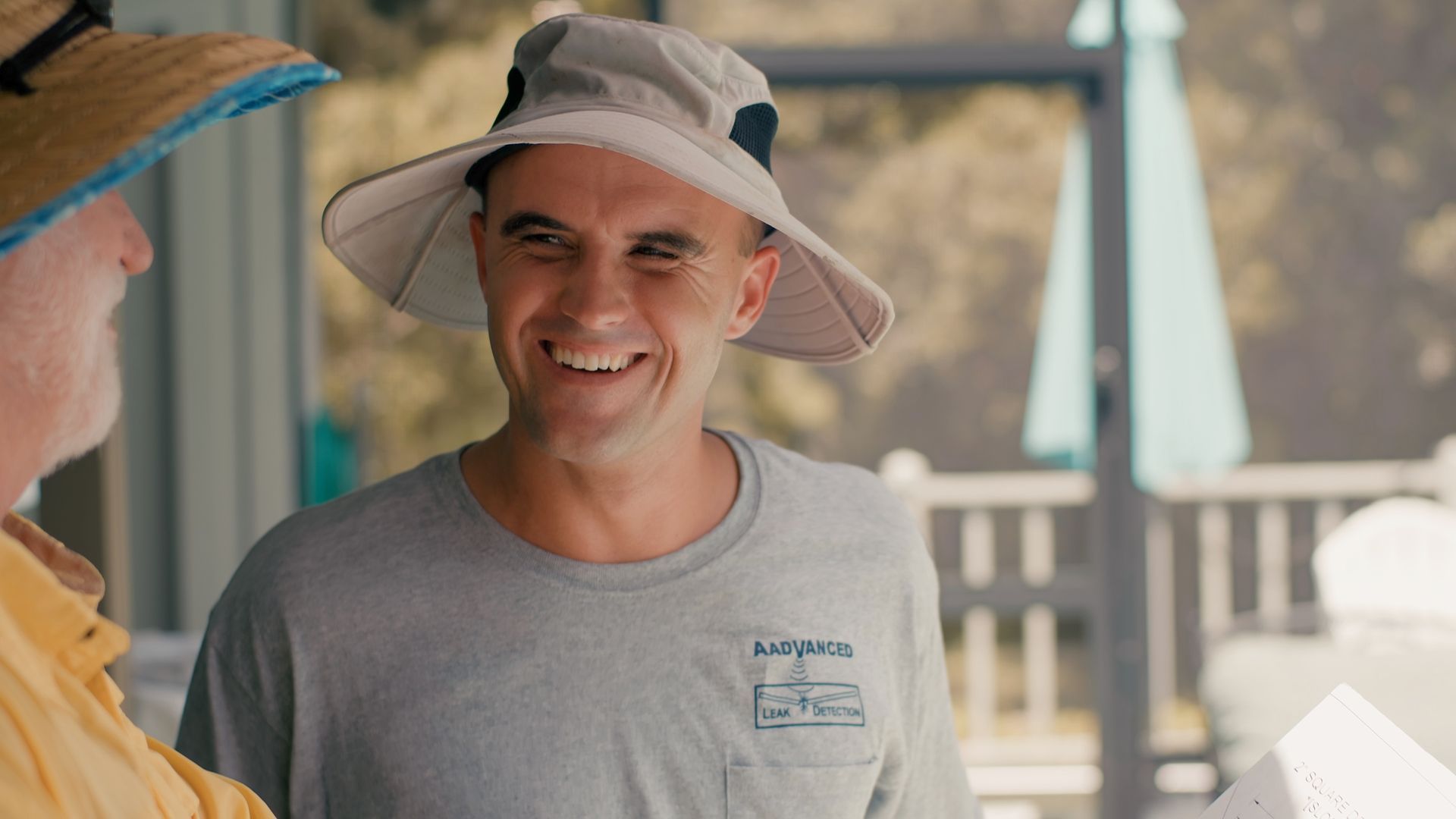 Man in sun hat smiles, standing outdoors next to a person.