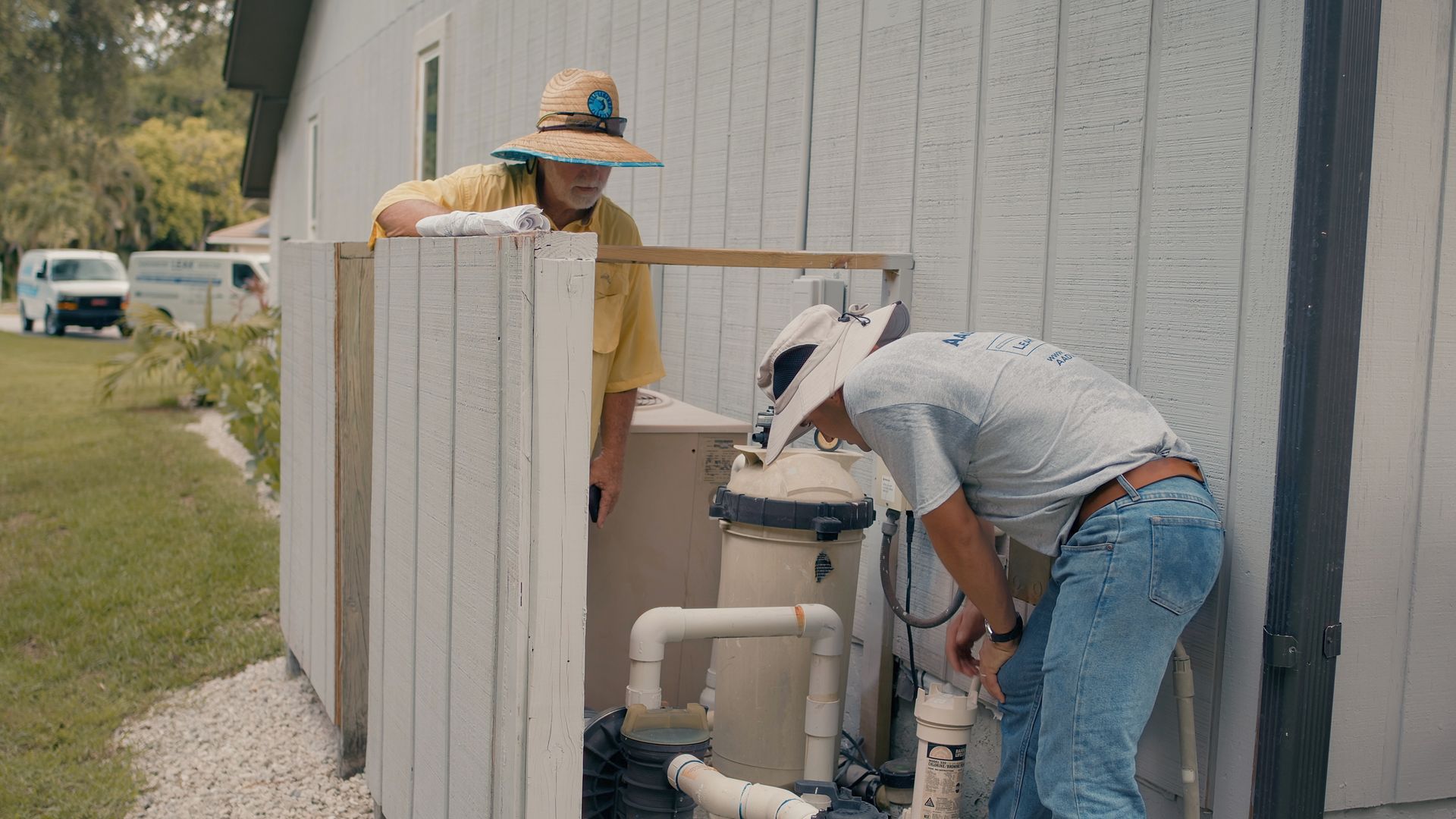 Two people working on a pool filter system next to a white building. One person in a yellow shirt and a straw hat observes.