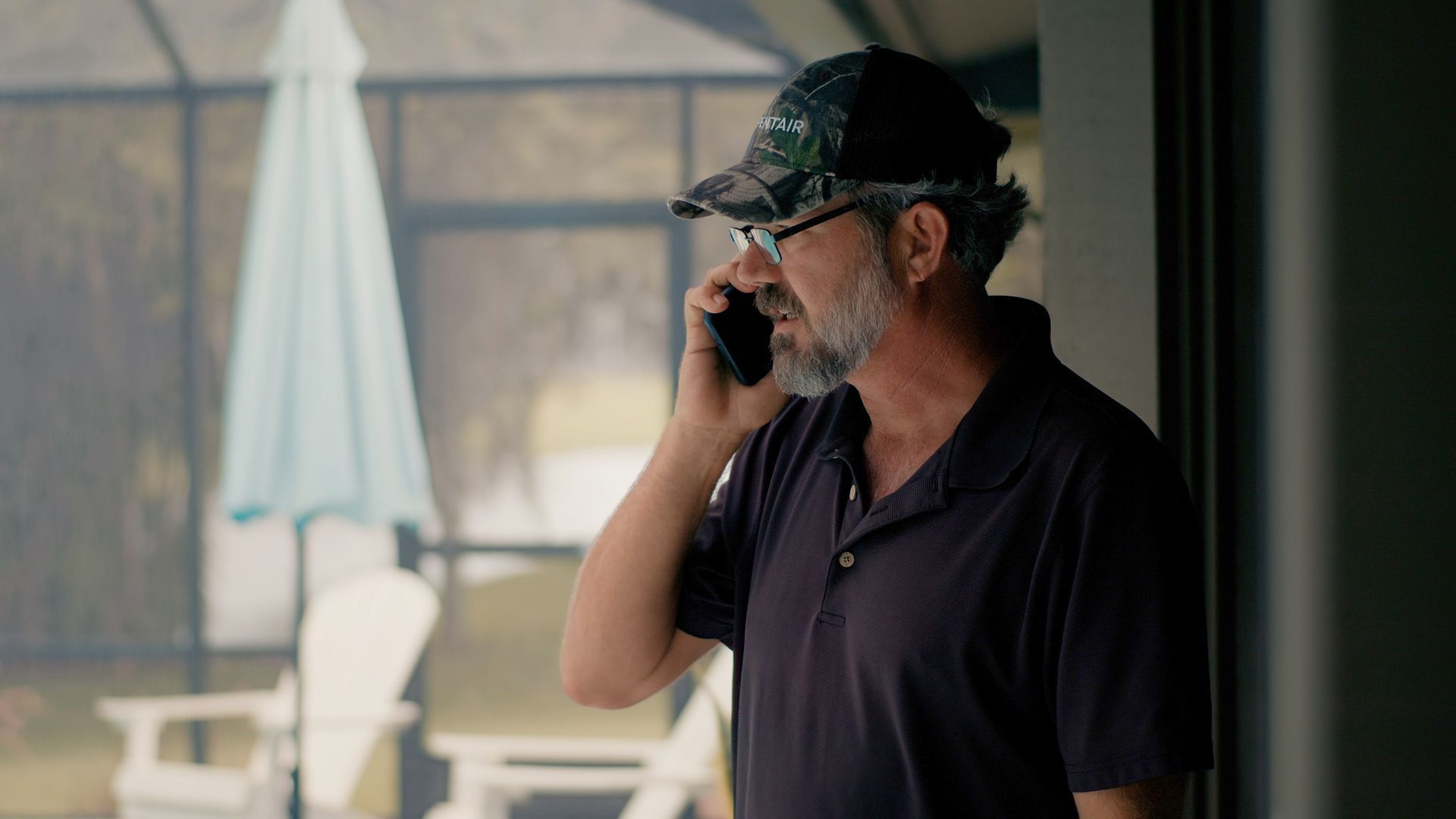 Man in camo cap and glasses on a phone, looking out a glass door towards a patio.