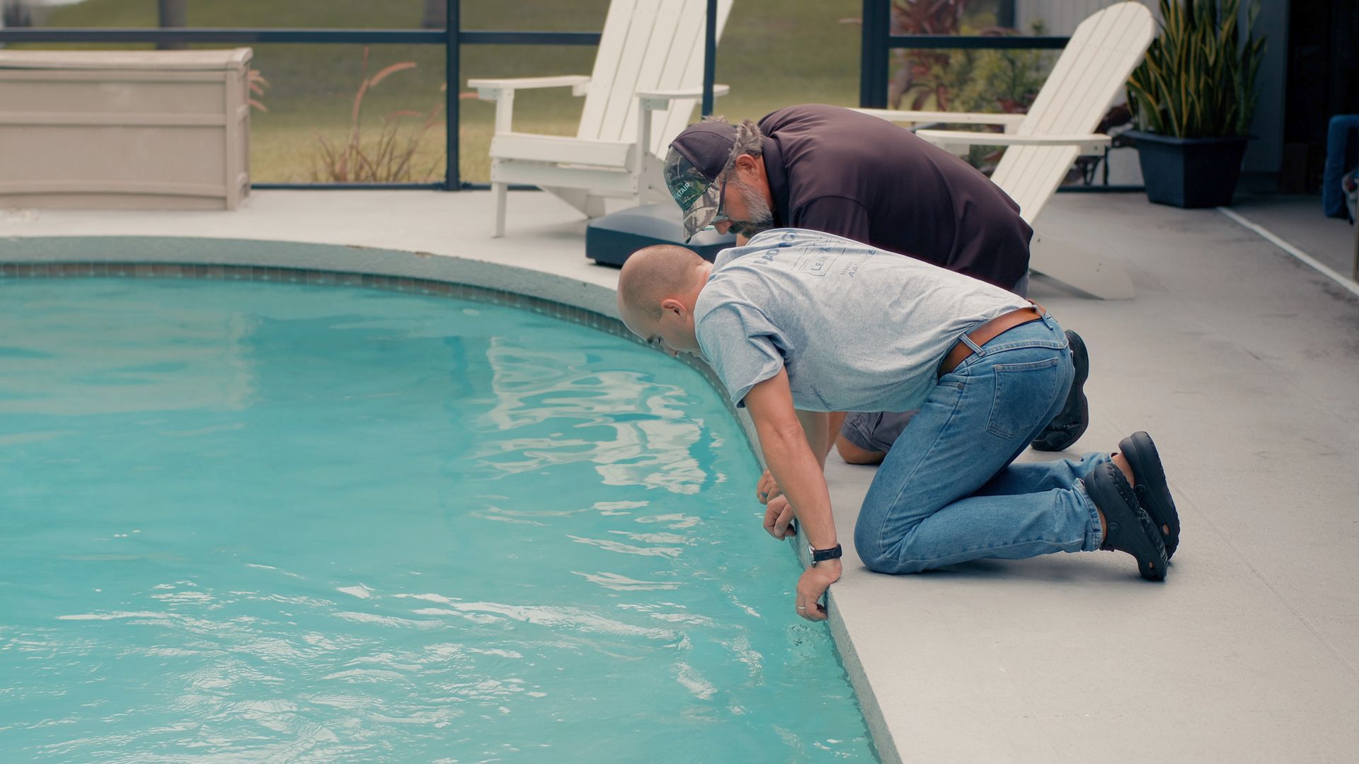 Two men inspecting a pool's water, kneeling by the edge. The pool is light blue, backyard setting.