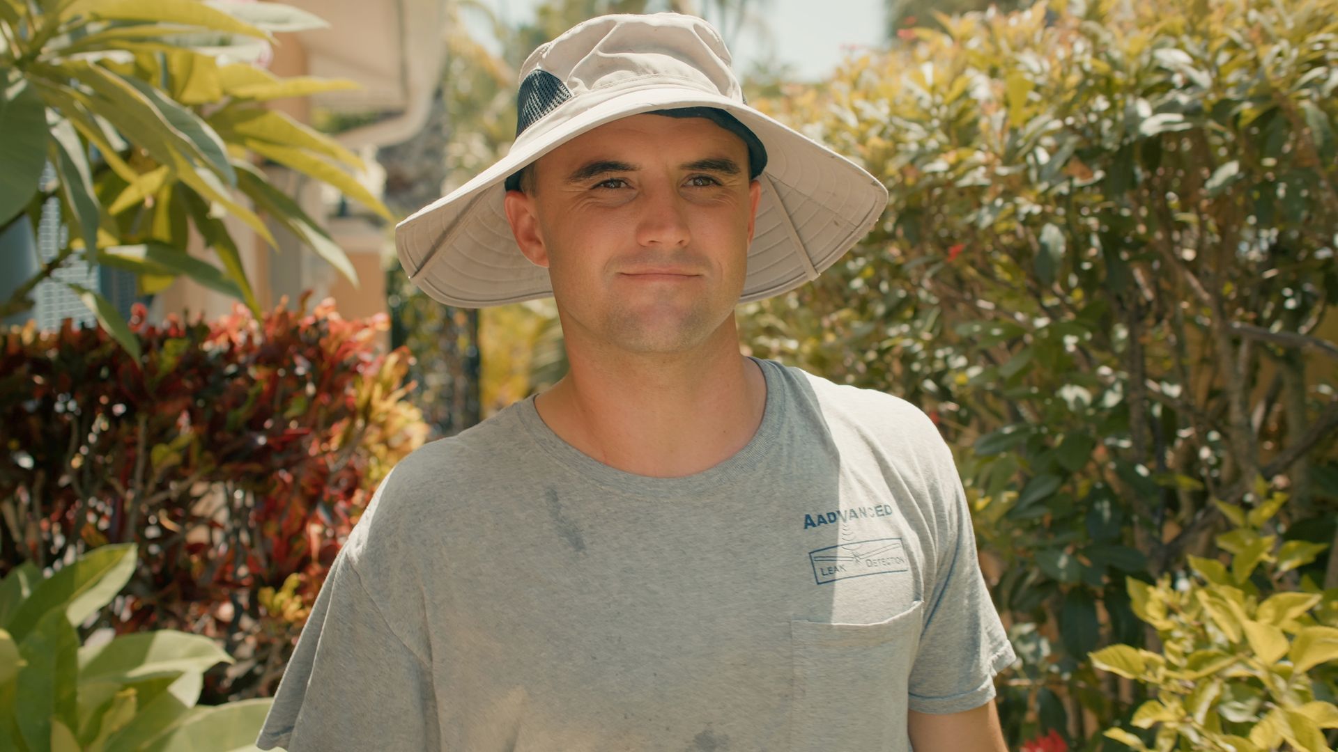 Man wearing a wide-brimmed hat and gray t-shirt standing outside, surrounded by greenery.