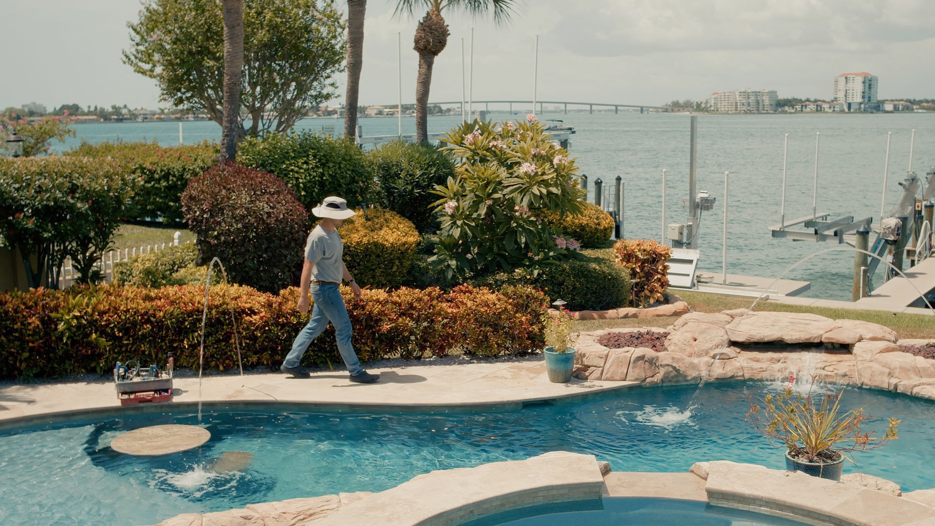 Man walks by a pool towards the waterfront, wearing a hat and jeans. Lush garden, blue water, sunny day.