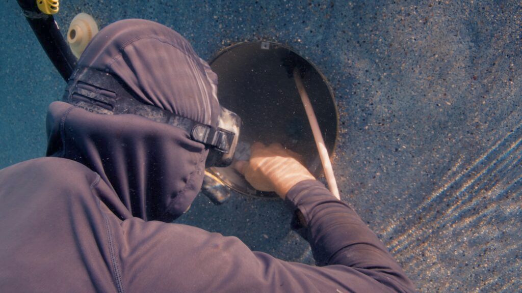 Person underwater, wearing diving gear, working on a dark, circular object with a tool. Water streams out.