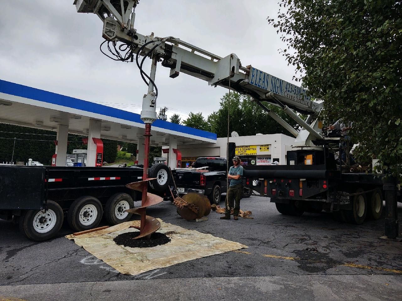A man is standing next to a truck with a crane attached to it in front of a gas station.