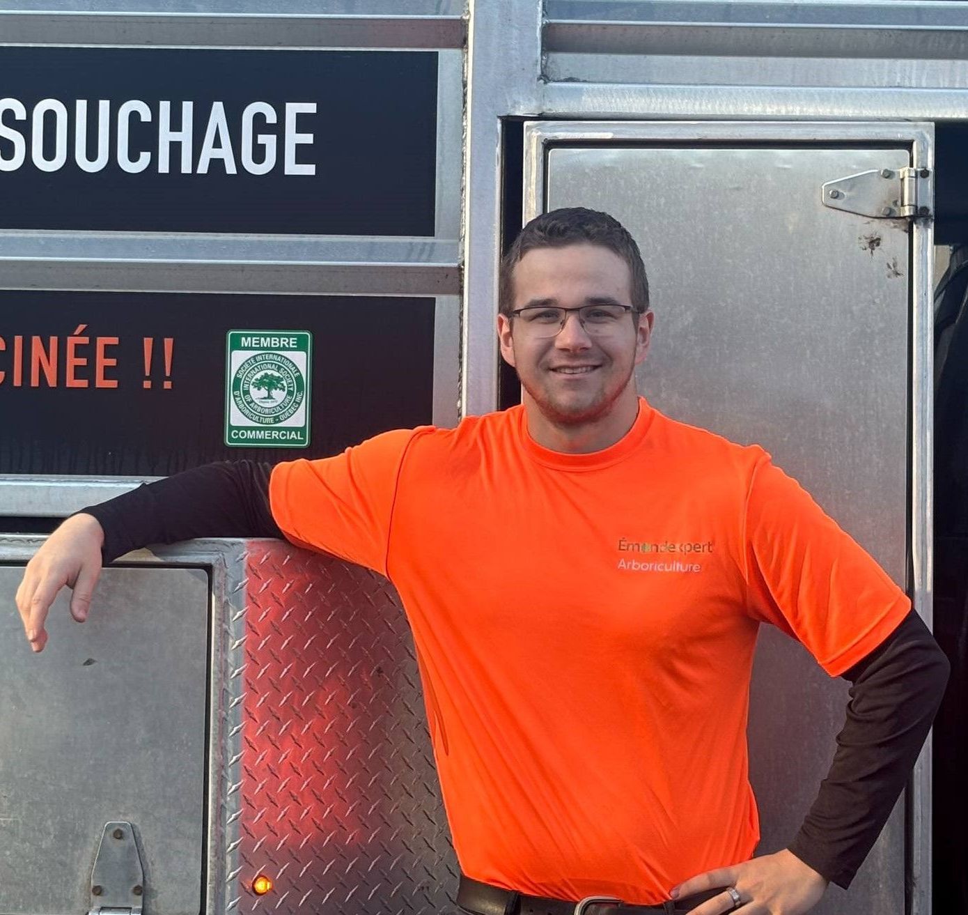 Un homme portant un gilet de sécurité et des lunettes de soleil se tient devant un panneau indiquant 50,025