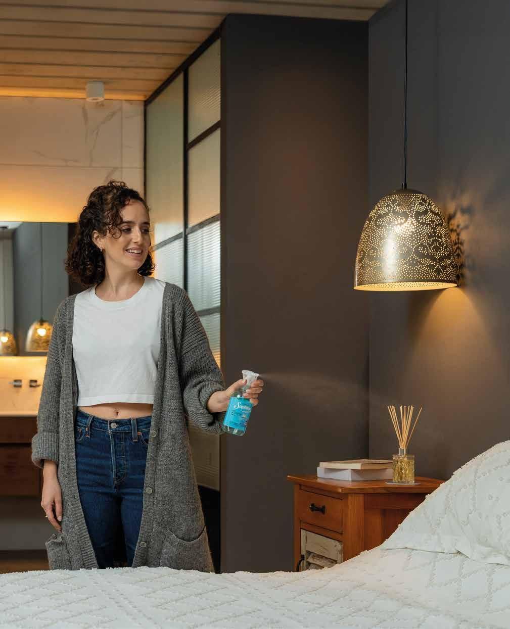 Mujer rociando ambientador en un dormitorio con muebles de madera, una lámpara colgante y un baño al fondo.