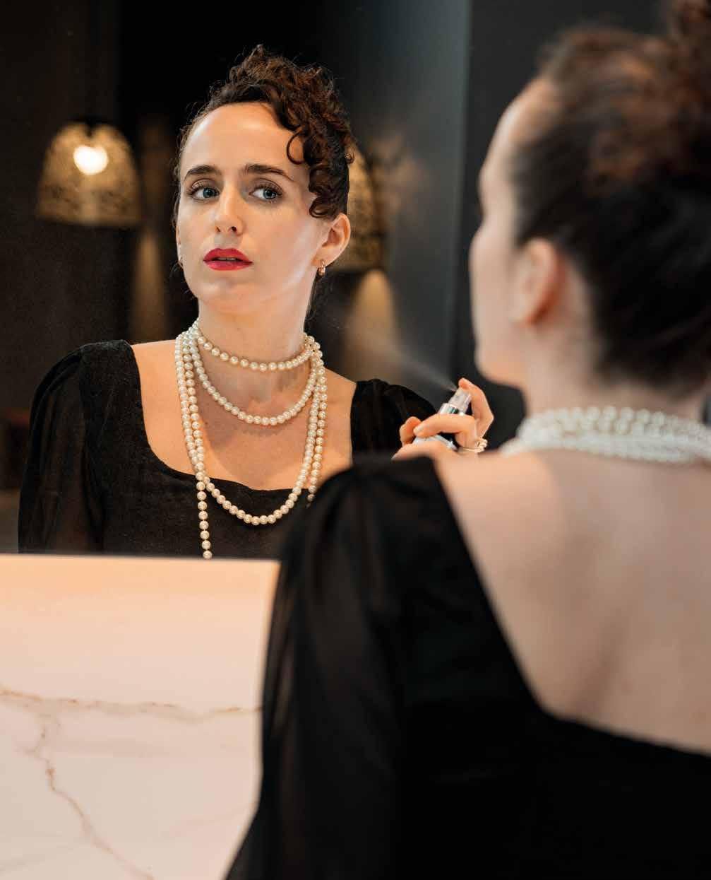Mujer con vestido negro y perlas se perfuma en el espejo. Lápiz labial rojo y cabello oscuro.
