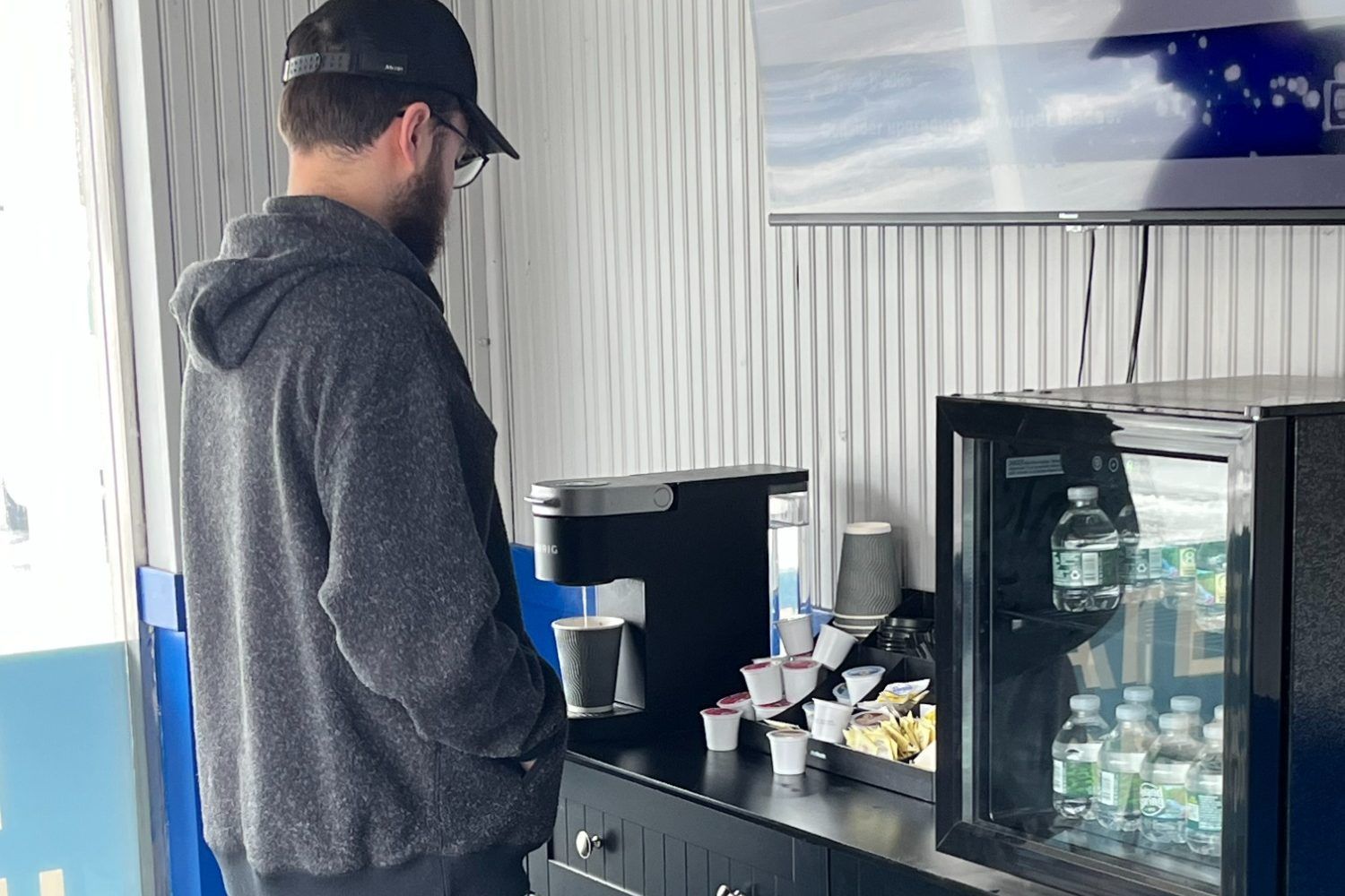Man in a grey hoodie and cap pours coffee from a machine. Coffee station with water bottles and cups in a waiting area | Diamond Automotive