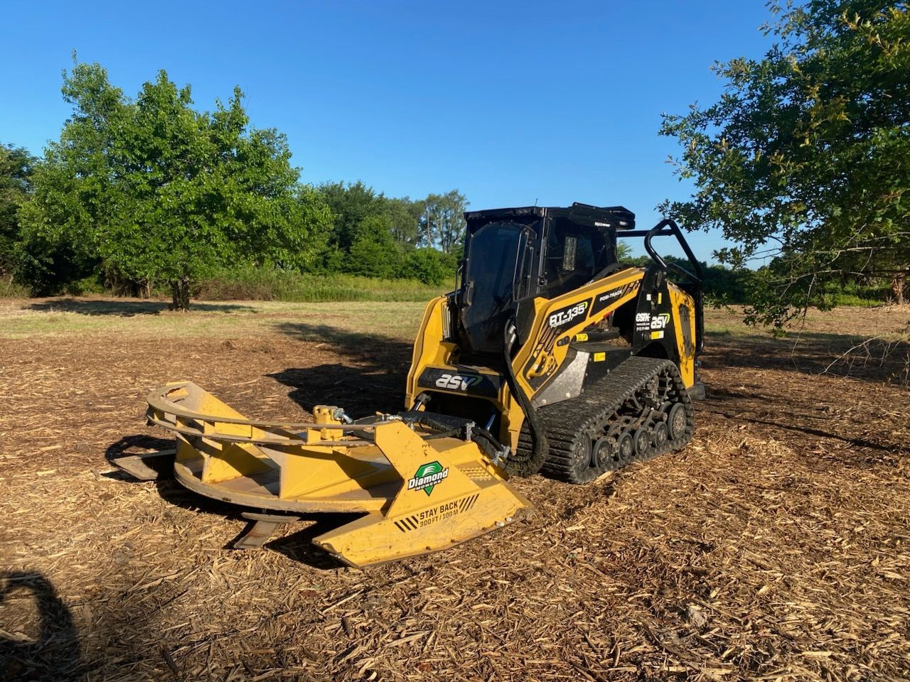 Yellow skid steer with mulching head clearing wood chips under trees on a sunny day.