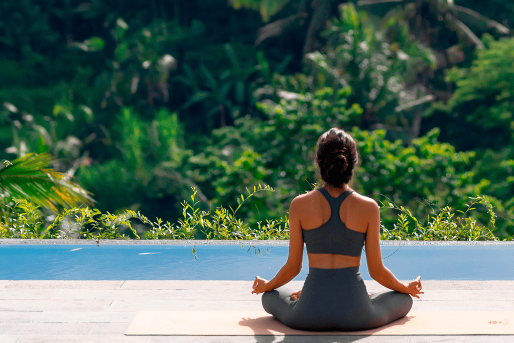 Texto alternativo - mulher de costas com as pernas cruzadas em posição de meditação. Ela usa um top e uma calça cinza. Ela está virada para um local aberto, com diversas árvores.