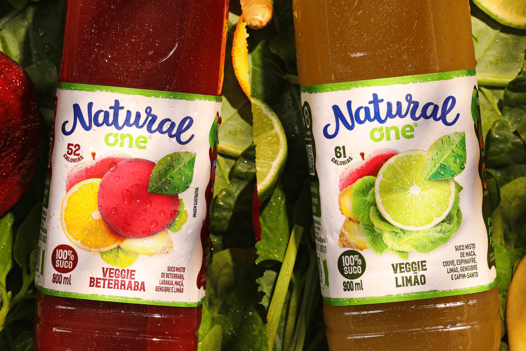 Texto alternativo - duas garrafas de suco Natural One. Do lado direito, há uma garrafa de veggie limão e ao lado esquerdo, uma garrafa de veggie beterraba. Ao fundo da imagem, há diversas hortaliças.